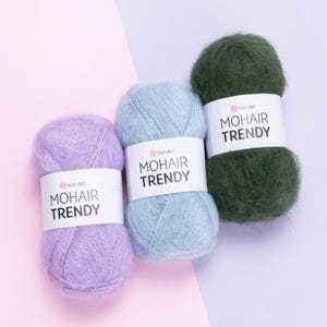 Può includere: Tre gomitoli di filato mohair nei toni lavanda, azzurro e verde scuro. Ogni gomitolo ha un'etichetta bianca con la scritta "MOHAIR TRENDY" e il logo "YamArt". Il filato è soffice e morbido, ideale per progetti di maglia o uncinetto.
