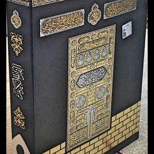 Op de afbeelding: Een zwarte decoratieve doos met gouden en zilveren versieringen, met sierlijke Arabische kalligrafie en een gedetailleerd deurontwerp. De doos heeft een baksteenpatroon aan de basis en een rood lint-bladwijzer.