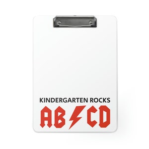 Könnte beinhalten: Ein weißer Clipboard mit einem schwarzen Clip und dem Text "Kindergarten Rocks" in schwarzen Buchstaben. Unter dem Text befindet sich ein roter Blitz zwischen den Buchstaben "AB" und "CD" in rot.