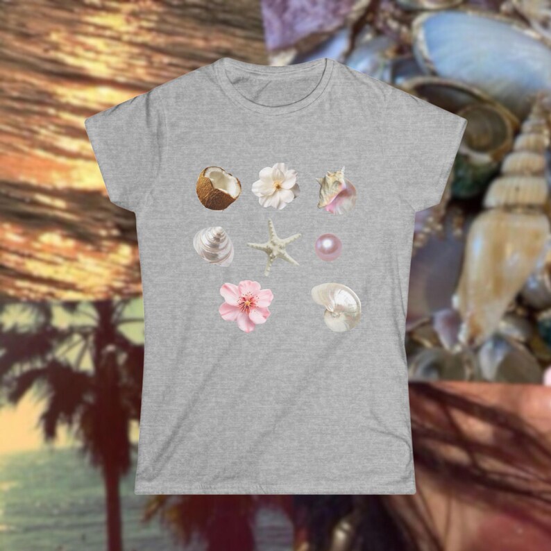 May include: A heather gray t-shirt featuring a collection of watercolor illustrations of seashells, flowers, a coconut, and a pearl. The design includes a starfish and a variety of shells in shades of white, pink, and brown.