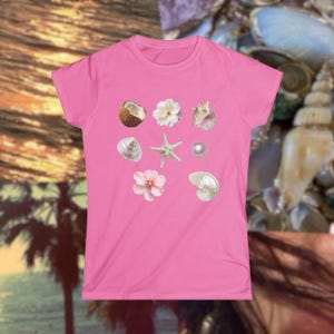 May include: A pink t-shirt featuring a collection of seashell and floral graphics. The design includes a coconut, white flowers, a seashell, a starfish, a pearl, and a pink flower. The shirt is a solid pink color.