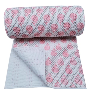 Op de afbeelding: Een opgerolde, witte quilt met een roze bloemenpatroon en grijze stippenaccenten. De quilt is gedeeltelijk uitgevouwen en onthult een gewatteerd interieur met roze stiksels. Het algehele ontwerp is een bloemenpatroon.