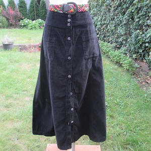 Op de afbeelding: Een zwarte midi-rok van corduroy met een knoopsluiting aan de voorzijde met donkere ronde knopen, twee opgestikte zakken aan de voorzijde en een hoge taille. De rok is gecombineerd met een gevlochten riem met een veelkleurig patroon.