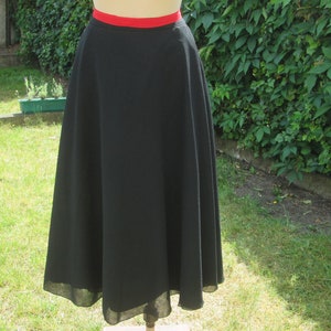 May include: A black skirt with a red waistband. The skirt is a full, flowing style, likely made of a lightweight fabric. The hemline falls below the knees. The skirt is displayed on a mannequin in a garden setting.