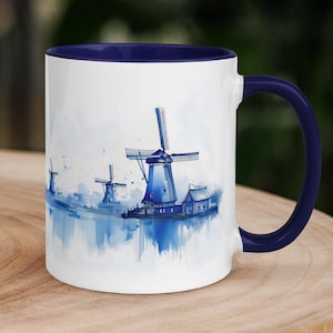 May include: White ceramic mug with a blue interior and handle. The mug features a watercolor painting of blue windmills on a white background. The design wraps around the mug, showcasing the windmills and their reflections.