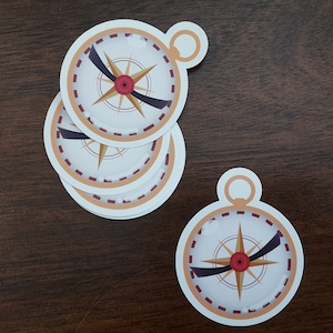 May include: Four white compass stickers with a gold compass design and a black needle. The compass has a red center and a gold ring around the edge. The stickers are on a brown wooden surface.