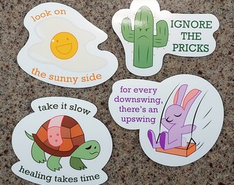 May include: Four colorful stickers with motivational messages. The first sticker features a sunny-side-up egg with the text "look on the sunny side". The second sticker features a green cactus with a grumpy face and the text "IGNORE THE PRICKS". The third sticker features a purple bunny on a swing with the text "for every downswing, there's an upswing". The fourth sticker features a turtle with the text "take it slow healing takes time".