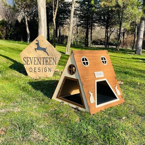 Puede incluir: Una casa de madera para conejos con un techo triangular y un exterior marrón. La casa tiene dos ventanas pequeñas y un letrero que dice "Bienvenido". La casa está sentada en un césped verde.