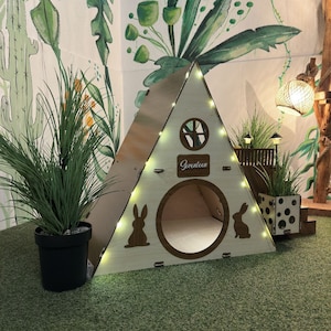 May include: A wooden triangular pet house with a round entrance, a small window, and the word "Seventeen" written on the front. The house is decorated with string lights and has a small wooden platform with a polka dot pattern.