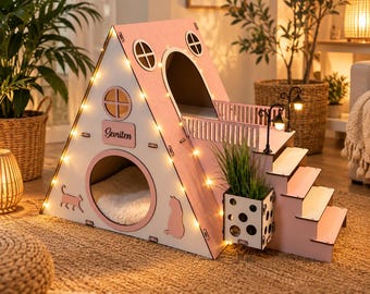 May include: A pink and white A-frame pet house with a circular entrance, a nameplate reading Sevriten, and a side staircase. The structure features decorative cutouts of cats, small windows, a balcony with two miniature lamps, and string lights.