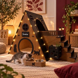 May include: A wooden A-frame pet house with black trim, circular windows, and string lights. It features a side staircase, a balcony with two small lamps, and a nameplate reading Seventeen. A wooden double bowl feeder sits in front.