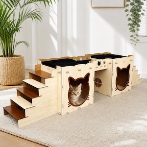 May include: A multi-level cat house made of light-colored wood. It features steps, cat-shaped cutouts, and a black cushioned resting area. A cat is visible inside one of the cutouts. The design includes a bridge and a circular opening.