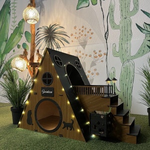 Puede incluir: Una casa para gatos de madera con forma de triángulo, con un techo negro y un pequeño balcón. La casa tiene una puerta para gatos, una ventana y un letrero que dice "Seventeen". La casa está decorada con luces de cadena y se encuentra sobre un fondo de pared pintada con un cactus y plantas tropicales.