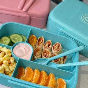 May include: A blue lunch box filled with food, including cucumber slices, mandarin orange segments, and rolled-up wraps. A small container holds pink dip. A pink lunch box with a spoon and fork is in the background.