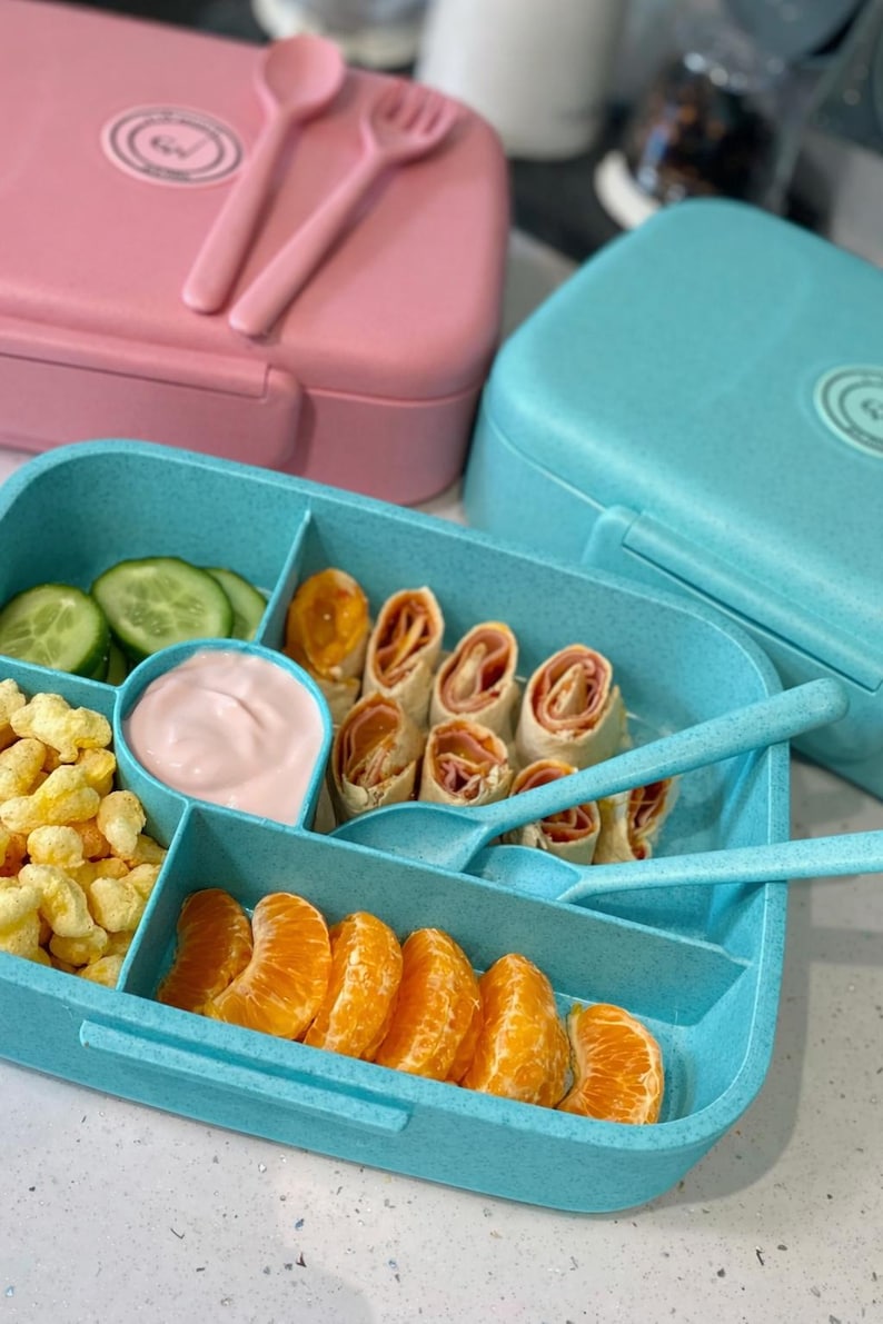 May include: A blue lunch box filled with food, including cucumber slices, mandarin orange segments, and rolled-up wraps. A small container holds pink dip. A pink lunch box with a spoon and fork is in the background.