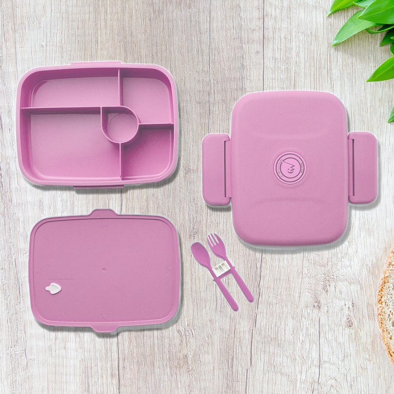 May include: A disassembled pink lunch box with a divided interior, lid, and matching fork and spoon set. The box is open, revealing the compartments. The lid has a circular emblem. The set is on a wooden surface.
