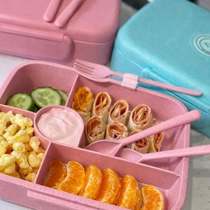 May include: Pink lunchbox with a divided tray filled with food, including cucumber slices, rolled wraps, and orange segments. The lunchbox includes a pink spoon and fork. A blue lunchbox is in the background.