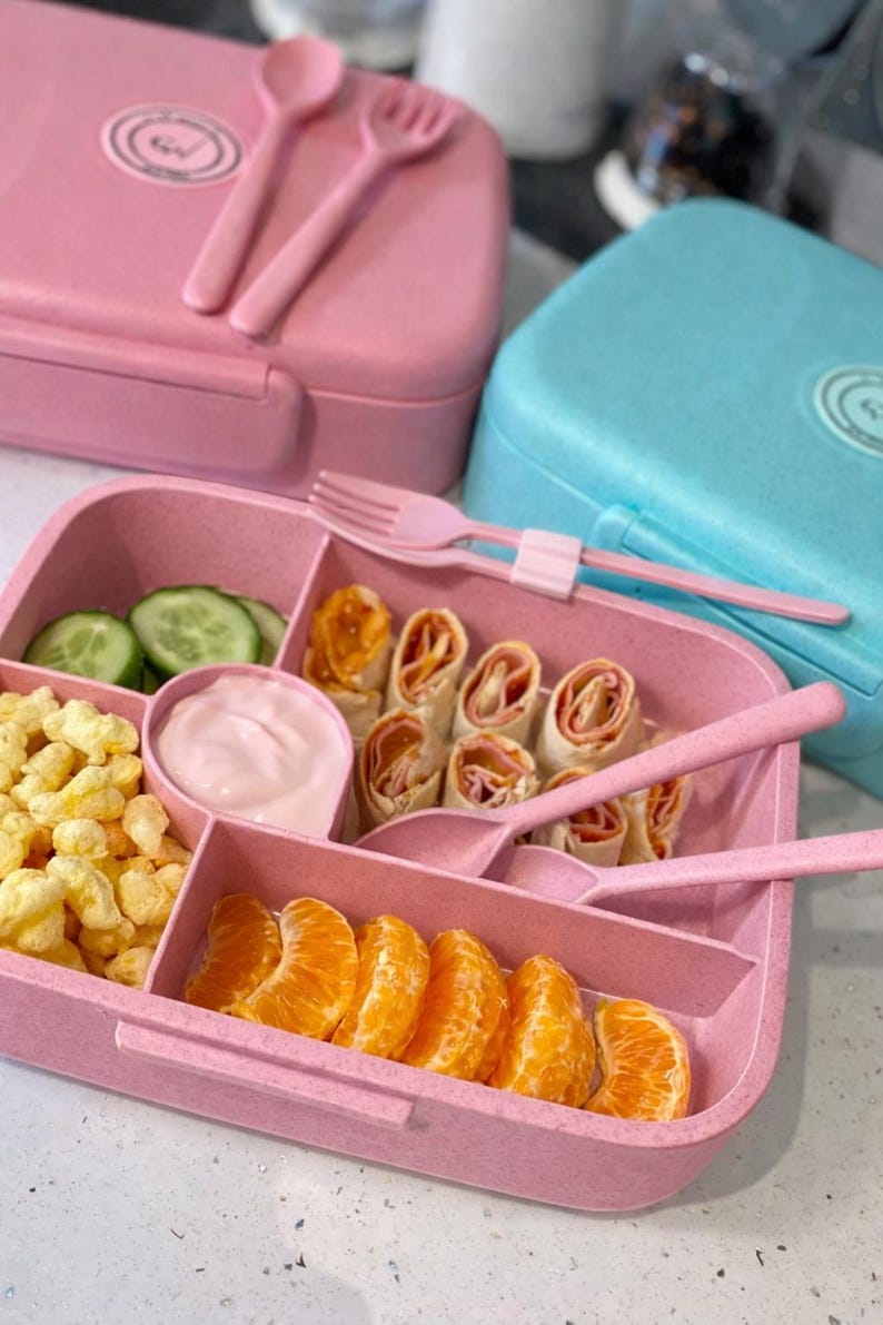 May include: Pink lunchbox with a divided tray filled with food, including cucumber slices, rolled wraps, and orange segments. The lunchbox includes a pink spoon and fork. A blue lunchbox is in the background.