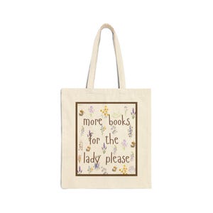 May include: A cream-colored canvas tote bag with two shoulder straps. The front features a rectangular brown border enclosing the brown text 'more books for the lady please' surrounded by small, colorful watercolor-style floral illustrations.