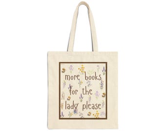 May include: A cream-colored canvas tote bag with two shoulder straps. The front features a rectangular brown border enclosing the brown text 'more books for the lady please' surrounded by small, colorful watercolor-style floral illustrations.