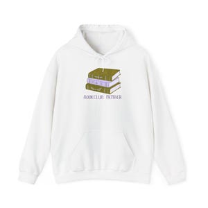 May include: A white hooded sweatshirt featuring a graphic of a stack of three books in olive green and light purple with floral details. Below the books, the text "BOOKCLUB MEMBER" is printed in a dark purple, sans-serif font.