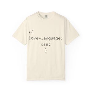 Può includere: Una t-shirt a maniche corte color crema con testo nero stampato sul davanti che recita: *{ love-language: css; }. La maglietta presenta un girocollo e una vestibilità standard, con un semplice design ispirato al codice informatico.