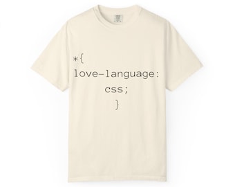 May include: A cream-colored short-sleeve t-shirt featuring black text printed on the front that reads: *{ love-language: css; }. The shirt has a crew neckline and a standard fit, showcasing a simple code-inspired graphic design.