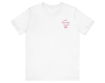 May include: A white short-sleeve t-shirt featuring a small pink pixelated graphic on the upper left chest. The graphic includes the text JUST ONE MORE LEVEL in a pixel font, flanked by two small star icons, with a pink game controller below.