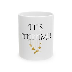 May include: A white ceramic mug featuring the black text IT'S TIIIIIIIME! with gold-colored hanging ornaments including gift boxes, a Christmas tree, a star, and a circular bauble dangling from the letters below the main phrase.