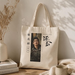 May include: Cream-colored canvas tote bag featuring a portrait of a person in a costume. The bag has black Chinese characters and the words "Chasing Jade" and "Zhang Linghe" printed on it. The bag has two handles.