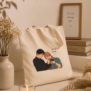 Peut inclure: Sac fourre-tout crème avec une illustration de deux personnes, dont une tenant un appareil photo. Le texte "Me & Thee" est imprimé sur le sac. Un cadre avec le texte "It was always you" est en arrière-plan.