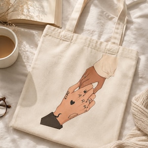 May include: A cream-coloured tote bag with an illustration of two hands holding each other. One hand has tattoos including a heart and the word "ARMY". The other hand wears a ring. The bag has two handles.