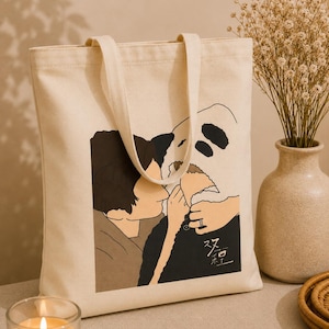 Peut inclure: Un sac fourre-tout en toile couleur crème arborant une illustration graphique d'une personne embrassant un panda. Le motif présente des tons noirs, marron et beige avec des caractères chinois imprimés dans le coin inférieur droit de l'image.