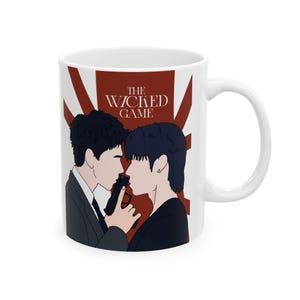 May include: White ceramic mug featuring a stylized illustration of two people facing each other, one holding a gun. The mug has the text "THE WICKED GAME" in red, with a red and white background.
