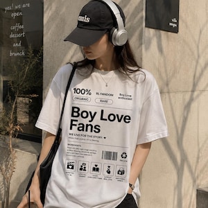 Peut inclure: T-shirt blanc avec le texte "Boy Love Fans" et du texte et des graphiques supplémentaires. Le t-shirt est porté avec une casquette noire et un casque. L'image est prise à l'extérieur près d'un café.