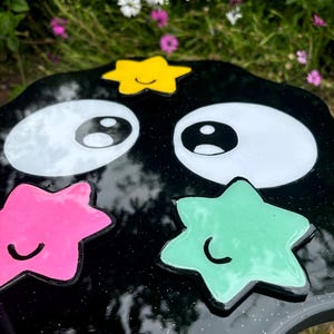 May include: Black resin table with a wavy edge and a whimsical design. It features two large white eyes, a yellow star with a smiley face, a pink star with a smiley face, and a green star with a smiley face.