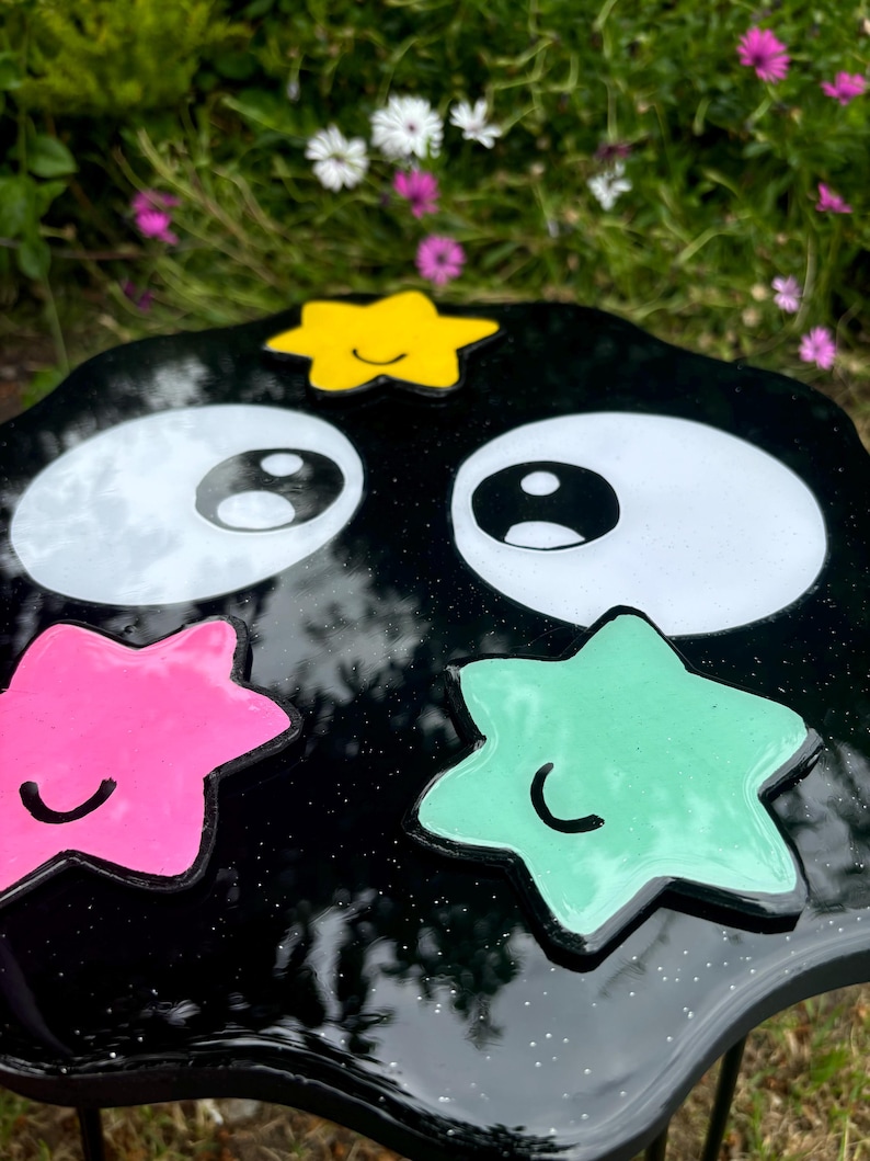 May include: Black resin table with a wavy edge and a whimsical design. It features two large white eyes, a yellow star with a smiley face, a pink star with a smiley face, and a green star with a smiley face.