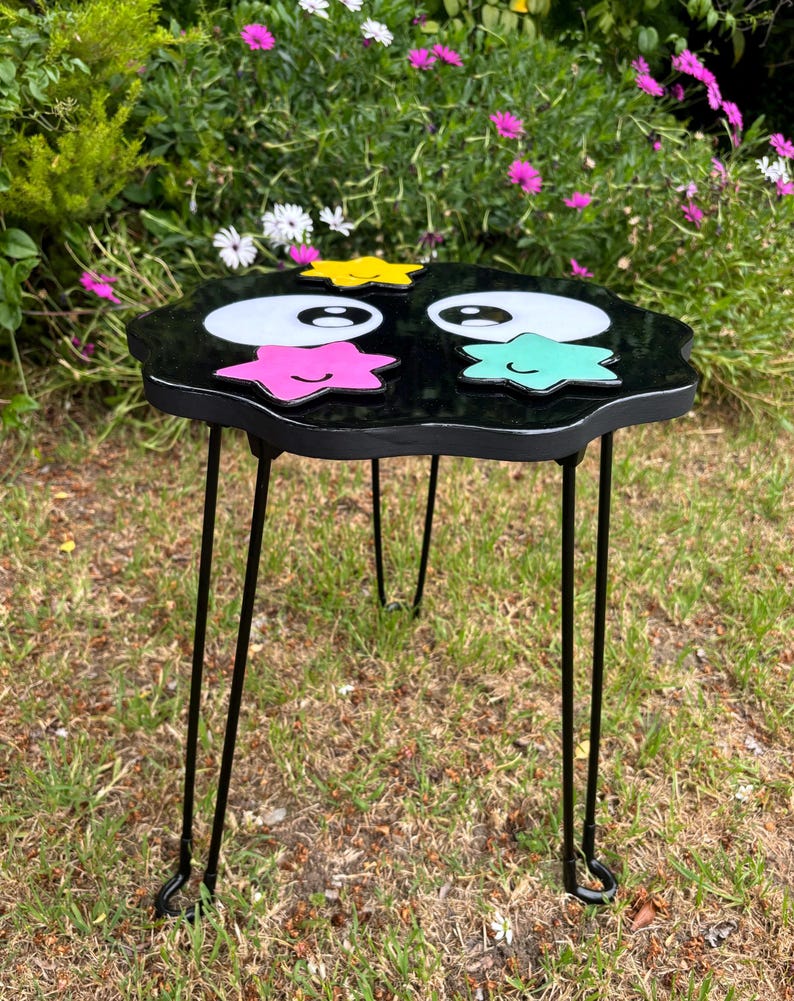 May include: Black wooden side table with a cloud-shaped top. The top is painted black and features two large white eyes with black pupils, a yellow star, a pink star with a smiling face, and a teal star with a smiling face. The table has three black metal legs.