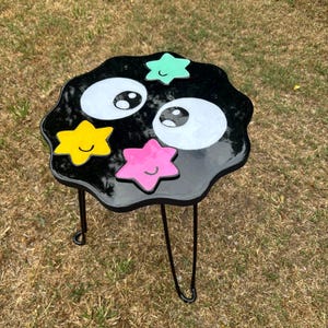 May include: Black side table with a black, white and green cartoon face and stars. The table has three black metal legs.