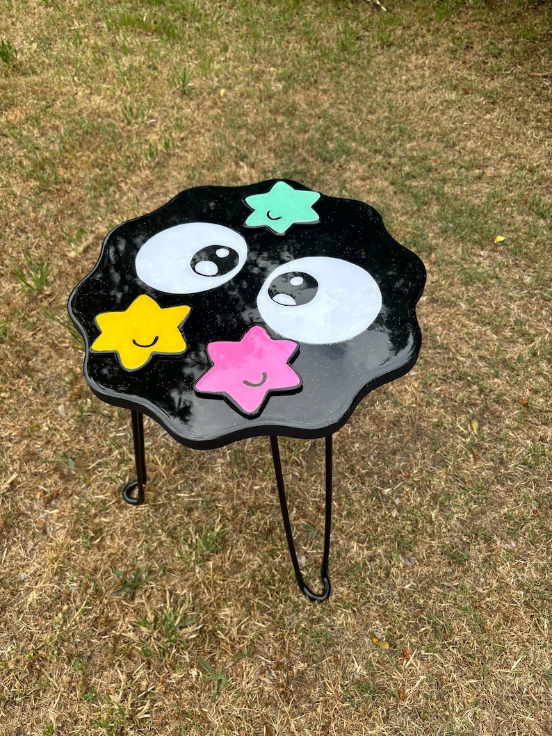 May include: Black side table with a black, white and green cartoon face and stars. The table has three black metal legs.