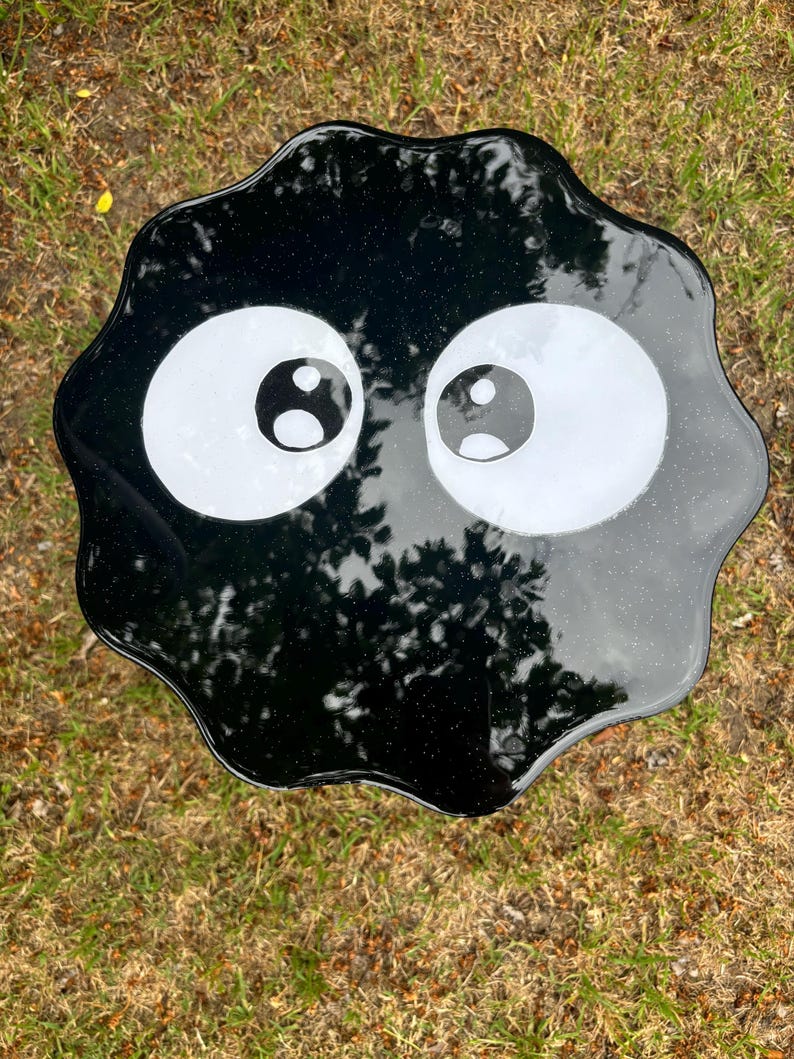 May include: Black resin table top with a wavy edge and two large white eyes with black pupils. The table top is reminiscent of a cartoon character.