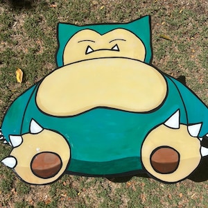 May include: A hand-painted wooden cutout of a green and brown cartoon character with white spikes. The character has a large, round belly and a wide, toothy grin.