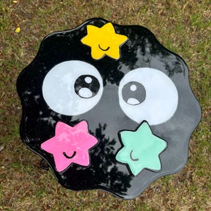May include: Black resin table with a wavy edge, featuring two large white eyes and four colourful stars with smiling faces. The stars are yellow, pink, green, and blue.