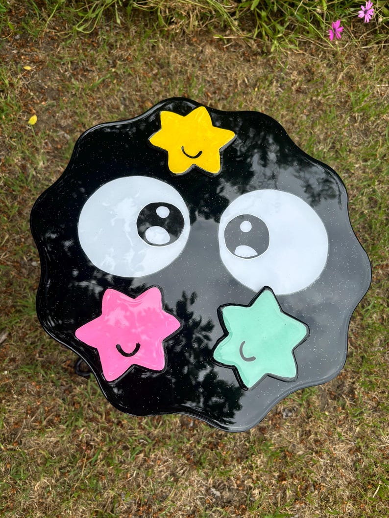 May include: Black resin table with a wavy edge, featuring two large white eyes and four colourful stars with smiling faces. The stars are yellow, pink, green, and blue.