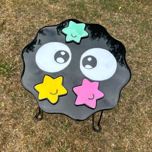 May include: Black and white resin table with a black metal frame. The table top is shaped like a cloud with two large eyes and four stars in different colours: green, yellow, pink, and blue. Each star has a smiley face.