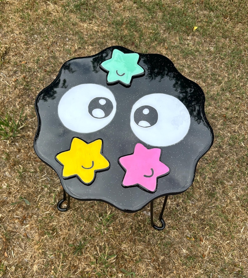 May include: Black and white resin table with a black metal frame. The table top is shaped like a cloud with two large eyes and four stars in different colours: green, yellow, pink, and blue. Each star has a smiley face.