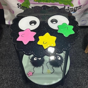 May include: A quirky black side table with a glittery top, cartoon eyes, and colourful star accents. The table has a light blue shelf with cartoon characters. The table is supported by black legs.
