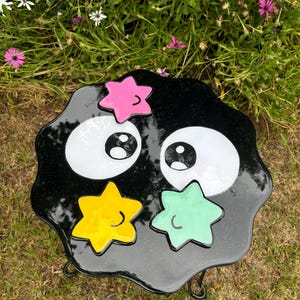 May include: Black and white cartoon-style table with large eyes and a smiling mouth. The table has a black, glittery surface with three yellow, green, and pink stars. The table is on a black metal stand with three legs.