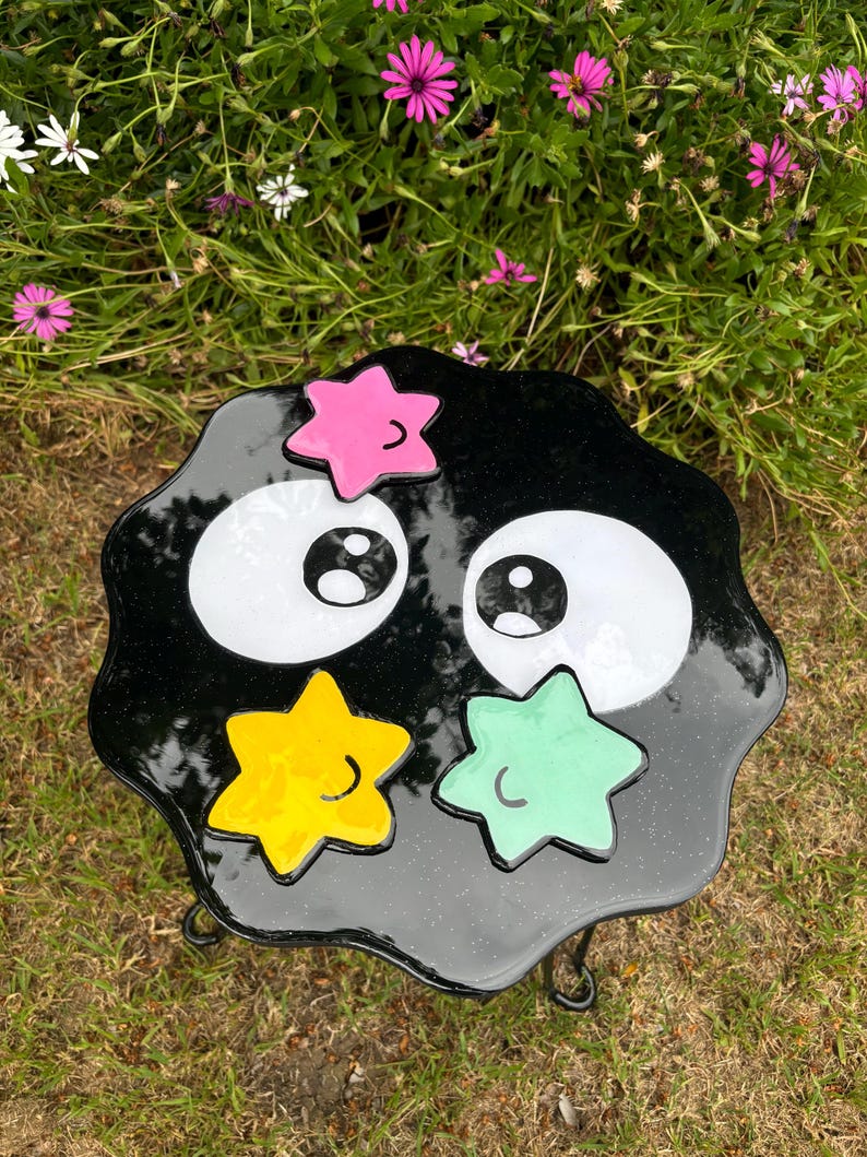 May include: Black and white cartoon-style table with large eyes and a smiling mouth. The table has a black, glittery surface with three yellow, green, and pink stars. The table is on a black metal stand with three legs.