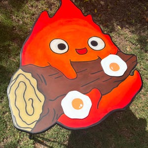 May include: A hand-painted wooden table shaped like a cartoon fire spirit holding a log with two fried eggs on top. The fire spirit is orange and red with a happy expression and large eyes. The log is brown with a yellow wood grain pattern.